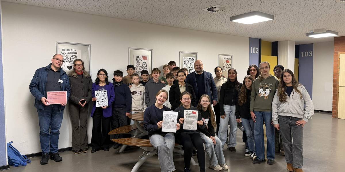 AES-Schulleiter Christian Augustin, Gleichstellungsbeauftragte Nicole Hendrych, Lehrerin Maria Ferndandéz, Bürgermeister Kai Eggert und Schülerinnen und Schüler einer siebten und zwölften Klasse eröffnen die Ausstellung „Red Flag – Bleib auf Augenhöhe“. © Stadt Laatzen Eine Gruppe aus Kindern, Jugendlichen und Erwachsenen steht in einem Schulflur vor Plakaten mit Comics.