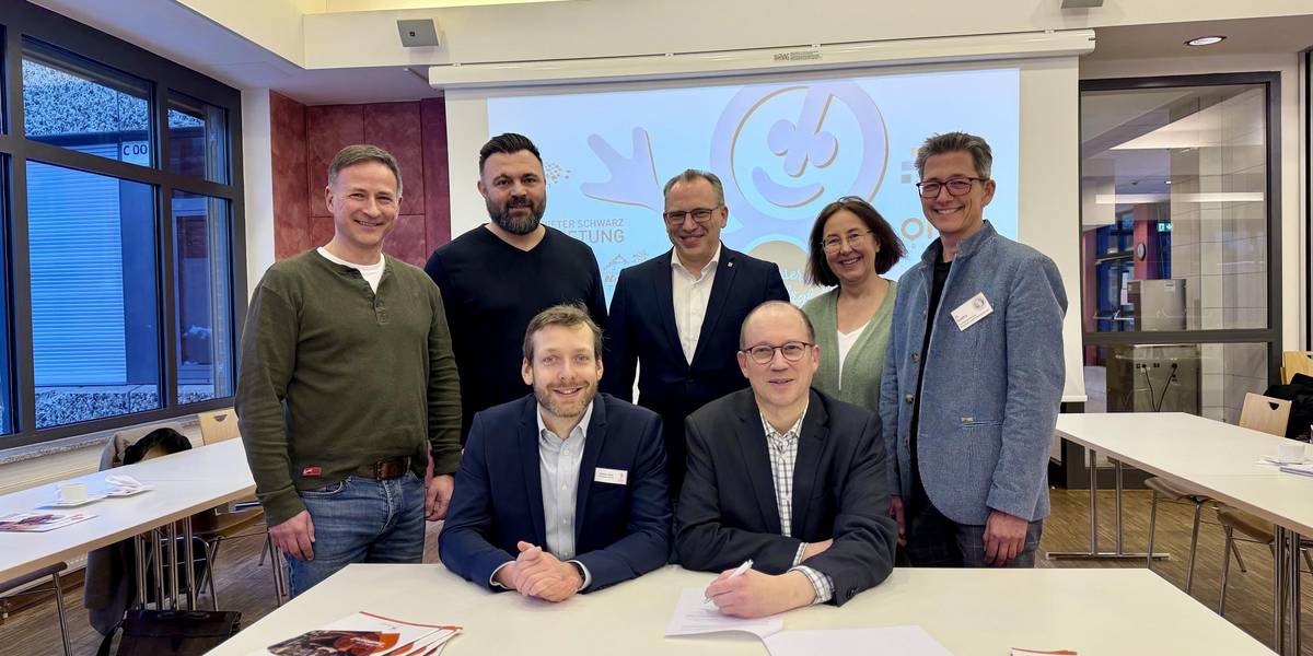 Zwei Männer sitzen an einem Tisch vor einer Projektion; hinter ihnen stehen fünf weitere Personen und schauen freundlich in die Kamera.