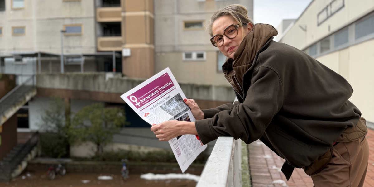 Eine Frau mit festem Blick lehnt über eine Brüstung, in der Hand hält sie eine Zeitung mit violettem Deckblatt.