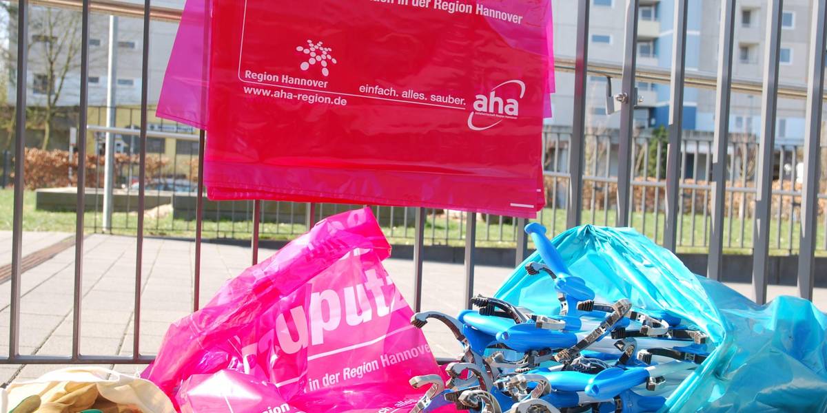 Einige rote Mülltüten und Müllzangen liegen an einem Metallzaun.