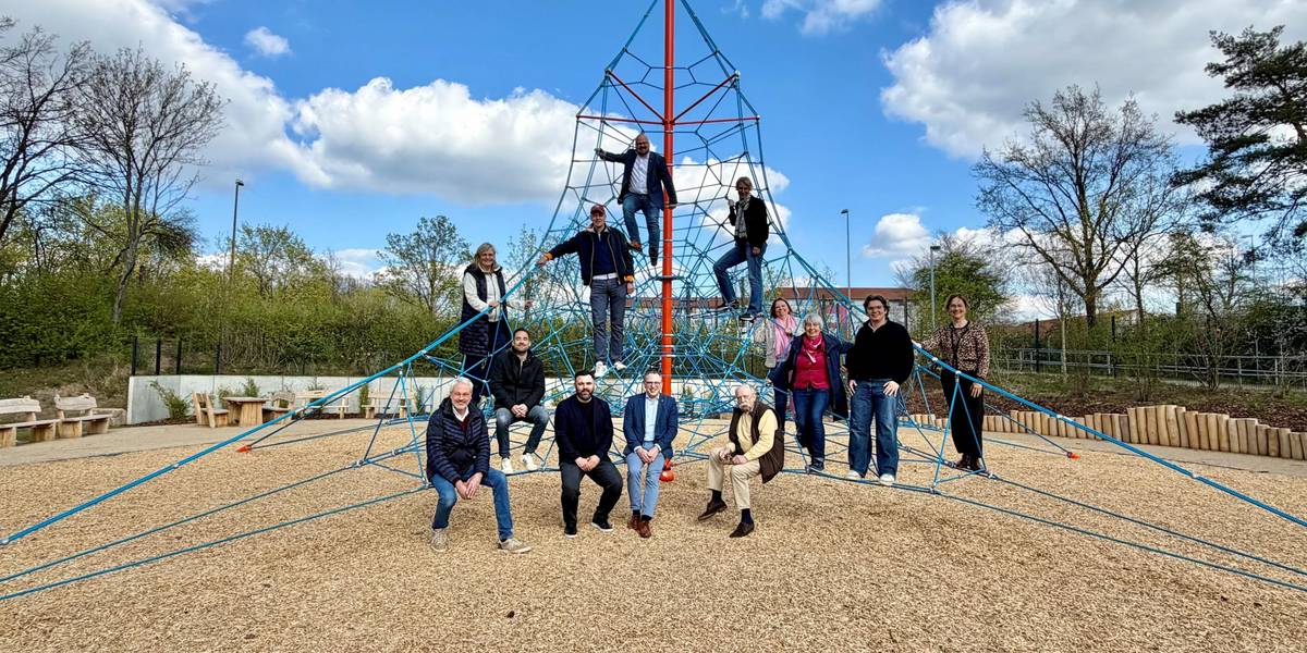 Eine Gruppe von 13 Personen hat eine Seilpyramide auf einem Spielplatz erklommen und sie schauen freundlich in die Kamera.