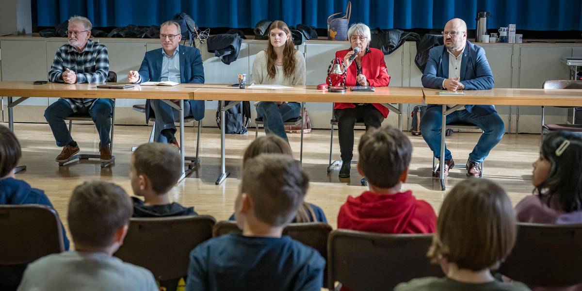 Ein Tisch, an dem mehrere Personen sitzen. Vor der Tischreihe befinden sich Kinder im Publikum. Von den Kindern sind nur die Rücken zu sehen.