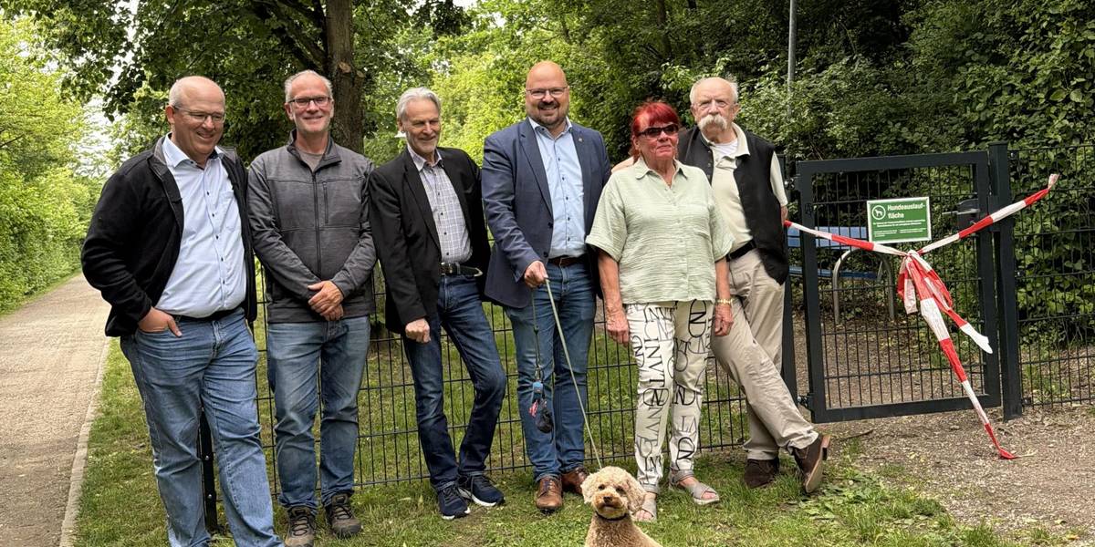 Eine Gruppe Männer und Frauen stehen mit einem Hund an der Leine vor einem eingezäunten Grundstück mit Wiese und Bäumen.