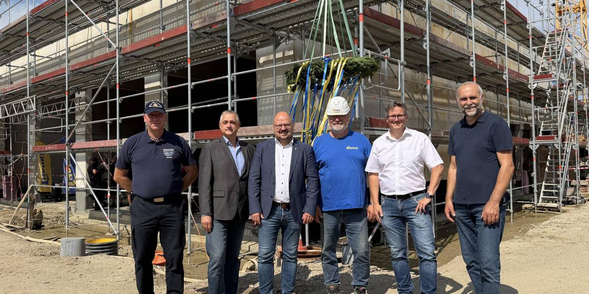 Drei Monate nach der Grundsteinlegung ist der Rohbau des neuen Feuerwehrhauses in Ingeln-Oesselse fertig. Beim Richtfest am 16. Juli 2026 feierten Ortsbrandmeister Frank Bezdiak (v.l.), Olaf Küpper, Geschäftsführer der Hanke Bau- und Projektentwicklungs GmbH, Bürgermeister Kai Eggert, Polier Markus Schmidt, Projektleiter Stefan Radke von Hanke Bau sowie Steffen Lauterbach von der arc architekturconzept GmbH den planmäßigen Baufortschritt. © Stadt Laatzen Eine Gruppe steht bei sonnigem unter einem Richtkranz.