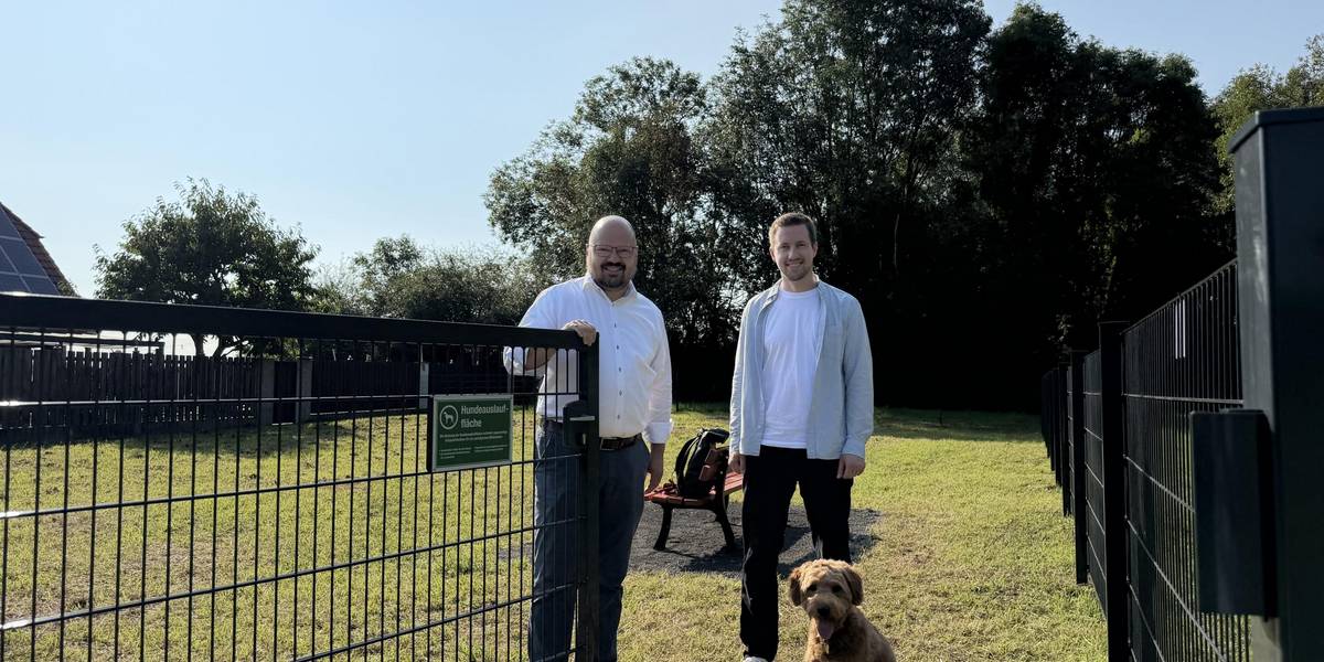 Platz zum Spielen und Toben: Bürgermeister Kai Eggert (l.) und Marvin Meyer, Leiter des städtischen Grünflächen-Teams, geben die über 500 Quadratmeter große Hundeauslauffläche am Sirupweg in Rethen offiziell frei. Hündin Maily wartet auf ihren Einsatz. © Stadt Laatzen Bürgermeister Kai Eggert und Leiter des städtischen Teams Grünflächen stehen an einem geöffneten Tor vor einer Hundewiese. Davor sitzt ein mittelgroßer Hund mit braunem Fell..