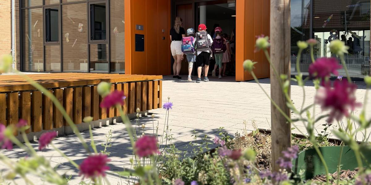 Grundschülerinnen und -schüler betreten mit Schulranzen auf dem Rücken einen orangenen Eingangsbereich einer Schule.