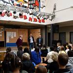 Zwei Männer stehen vor zahlreichen Kindern, die auf Stühlen sitzen.