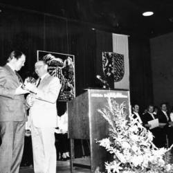 Landrat Jürgen Bauermeister übergibt ein Geschenk an einen Herrn, im Hintergrund sitzende Menschen und Fahnen an der Wand.