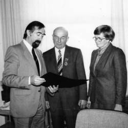 Ehrung mit zwei Herren mit Anzug und Krawatte sowie einer Dame mit Brille.