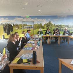 Die Teilnehmenden sitzen in U- Form am Tisch  und hören der Referentin zu.