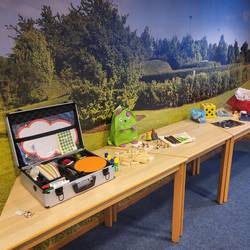 Es stehen verschiedene Spielmaterialien auf drei Tischen. Es sind Würfen, Spielfiguren, Aufkleber, Autos, Bausteine,...