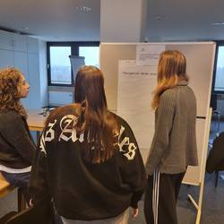 Drei FSL-lerinnen tauschen sich vor einer Metaplanwand zu einer Fragestellung aus. Die Ergebnisse schreiben sie auf ein Flipchartblatt, das an einer Metaplanwand hängt.