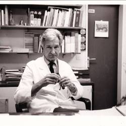 Mann im weißen Kittel sitzt am Schreibtisch, im Hintergrund Bücher und Tür