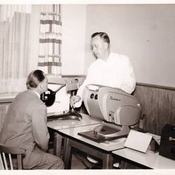 Herr macht einen Sehtest, hinter einem Tisch steht ein Herr mit weißem Kittel