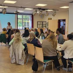 Die Teilnehmenden sitzen an einem langen Tisch. Sie hören der Referentin zu.