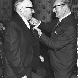 Ein Herr im Anzug steckt einem anderen eine Stecknadel an