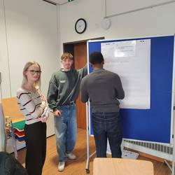 Eine Dreiergruppe steht vor einer Metaplanwand. Sie bearbeiten dort eine Fragestellung. Die Ergebnisse werden auf ein Flipchart geschrieben.