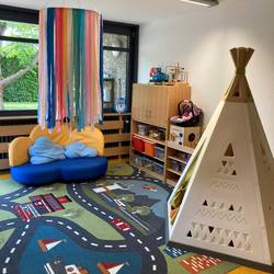 GTP Wichtelhöhle Gruppenraum © Stadt Laatzen Auf dem Boden liegt ein Spielteppich mit einer Straße drauf. Von der Decke hängt ein gebastelter Kranz mit langen Bändern. Dahinter an der Wand steht ein Sofa. Rechts an der Wand stehen Schränke mit Spielzeug. Rechts daneben ein Zelt.