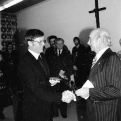 Zwei Herren im Vordergrund geben sich die Hand, im Hintergrund weitere Personen und ein Kreuz an der Wand.