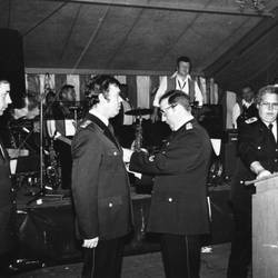 Vier Herren in Feuerwehruniform, einer davon vor Rednerpult, im Hintergrund Bühne mit Instrumenten