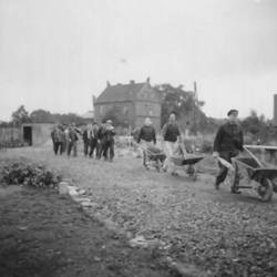 Männer mit Schubkarren auf Deldweg, im Hintergrund freistehendes Gebäude