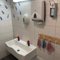 GTP Wichtelhöhle Badezimmer © Stadt Laatzen Im Badezimmer hängt auf Kinderhöhe ein Waschbecken mit bunten Wasserhähnen. Rechts neben dem Waschbecken hängt eine Leiste mit Haken für Waschlappen. Über dem Waschbecken hängt eine Lampe, ein Seifenspender, Desinfektionsspender und Papiertücher. Unter dem Waschbecken steht ein Mülleimer. In der linken Ecke an der Wand sind Wassertropfen aufgeklebt.