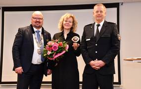 Laatzens Bürgermeister Kai Eggert, Corinna Luedtke und Leiter des Polizeikommissariats Laatzen stehen auf einer Bühne. Corinna Luedtke hält den Couragepreis in Form eines Auges sowie einen Blumenstrauß in den Händen. Alle drei lächeln in die Kamera.