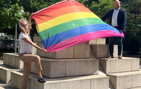 Ein Mann und eine Frau stehen auf Steinstufen. Zwischen ihnen weht eine Regenbogenflagge, die beide in ihren Händen halten.