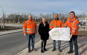 Drei Männer in orangenen Warnjacken und eine Frau stehen an der Einmündung einer neuen Straße auf dem Gehweg.
