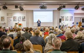 Blick über sitzende Menschen auf einen Redner an einem Rednerpult auf einer Bühne.