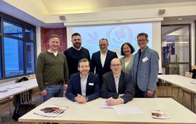 Zwei Männer sitzen an einem Tisch vor einer Projektion; hinter ihnen stehen fünf weitere Personen und schauen freundlich in die Kamera.