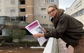 Eine Frau mit festem Blick lehnt über eine Brüstung, in der Hand hält sie eine Zeitung mit violettem Deckblatt.