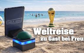 Tablet und Buzzer stehen am Strand vor dem Meer, im Hintergrund sind Menschen im Meer, ein Volleyball und der WM Pokal stehen am Strand