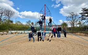 Eine Gruppe von 13 Personen hat eine Seilpyramide auf einem Spielplatz erklommen und sie schauen freundlich in die Kamera.