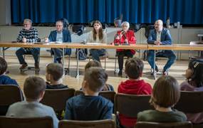 Ein Tisch, an dem mehrere Personen sitzen. Vor der Tischreihe befinden sich Kinder im Publikum. Von den Kindern sind nur die Rücken zu sehen.