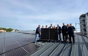 Eine Gruppe steht auf einer Dachfläche mit zahlreichen PV-Modulen und hält ein Solarpaneel in die Kamera.