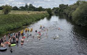 Schwimmerinnen und Schwimmer befinden sich in einem Fluss. Sie sind von oben, von einer Brücke aus fotografiert worden.