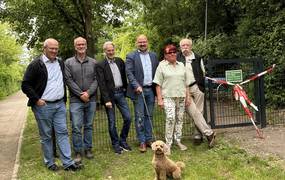 Eine Gruppe Männer und Frauen stehen mit einem Hund an der Leine vor einem eingezäunten Grundstück mit Wiese und Bäumen.