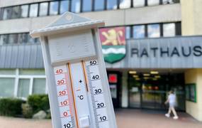 Eine Hand hält ein analogen Thermometer; im Hintergrund sieht man das Rathaus
