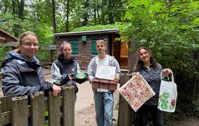 Drei Frauen und ein Mann stehen an einer Hütte im Wald und halten verschiedene Bastelwerke in die Kamera