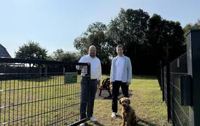Bürgermeister Kai Eggert und Leiter des städtischen Teams Grünflächen stehen an einem geöffneten Tor vor einer Hundewiese. Davor sitzt ein mittelgroßer Hund mit braunem Fell..