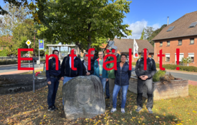 Eine Gruppe von Menschen stehen gemeinsam mit Bürgermeister Kai Eggert in Ingeln-Oessels vor einem großen Stein mit Gravur. Darüber der Schriftzug entfällt.