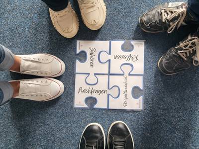 Auf dem Boden liegen 4 Puzzelteile. Auf den Puzzelteilen stehen Begriffe. Die Begriffe lauten Zuhören, Reflexion, Nachfragen und Perspektivwechsel.