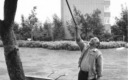 Mann zeigt mit Stab Richtung Baum, dahinter Schubkarre und Gießkanne auf einer Grünfläche, im Hintergrund ein Haus