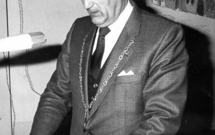 Mann mit Kette am Mikrofon, im Hintergrund ein Bild an der Wand