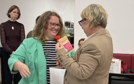 zwei Frauen stehen sich gegebüber. Die eine Frau überreicht das Zertifikat, ein Dokument, an die andere Frau.