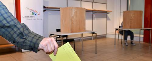 Eine Hand hält einen Wahlschein. Dieser Schein soll in die Wahlurne, die vor ihm steht.
