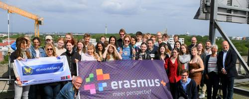 Eine große Gruppe junger Menschen steht auf einem Hochhausdach und lacht in die Kamera.