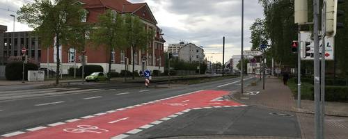 Roter Fahrradstreifen auf einer Straße. Rechts davon sind Geschäfte zu sehen.