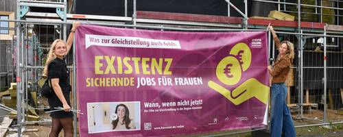 Zwei Frauen bringen ein Banner an einem Baustellenzaun an.